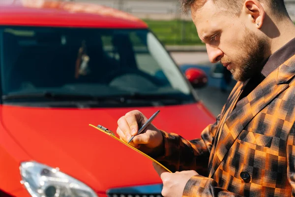 A man fills out a form document after an accident. Young guy European records data against the background of a red car