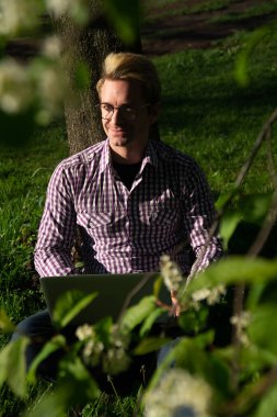 Ağacın altında oturup dizüstü bilgisayarda yazan beyaz bir adam. İnternet konsepti. Freelancer parkta çalışıyor. Güneşli bir günde. Çimenlerde boşluk kopyala