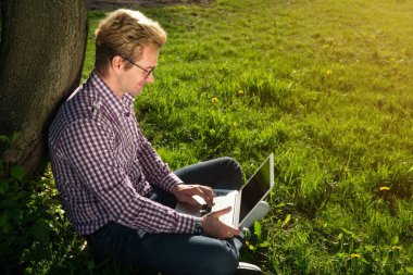Ağacın altında oturup dizüstü bilgisayarda yazan beyaz bir adam. İnternet konsepti. Freelancer parkta çalışıyor. Güneşli bir günde. Çimenlerde boşluk kopyala