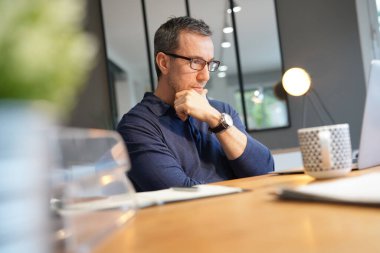 Orta yaşlı bir adam sıcak bir şeyler içiyor, dizüstü bilgisayarla çalışıyor.