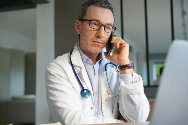 医者はオフィスでノートパソコンで電話で話してる — ストック写真