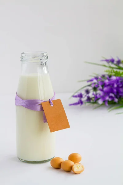 Garrafa de leite com etiqueta para escrever desejos, ao lado de seus doces de leite em fundo claro cinza de madeira e flores lilás do lado direito — Fotografia de Stock