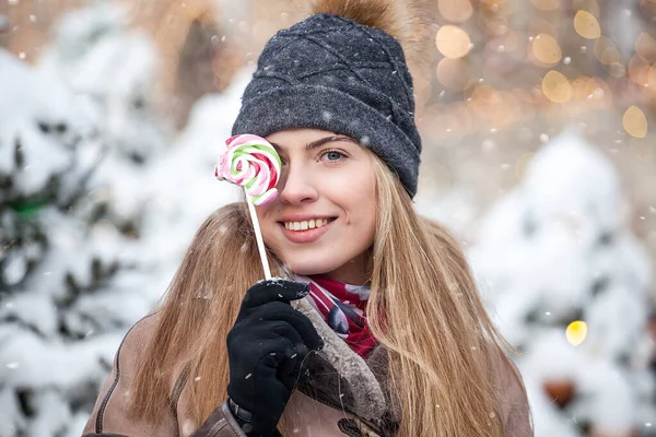 Smiling young woman dressed in winter clothes eating colorful Lollipop on Christmas snowy street — Stock Photo, Image