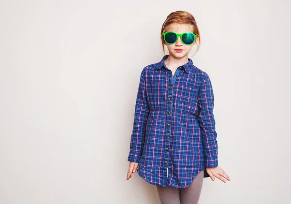 Redhead little girl in green sunglasses posing against wall. — Stock Photo, Image