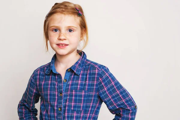Malá holčička zrzka v modré kostkované košili vystupují proti zdi. — Stock fotografie