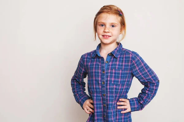 Little redhead girl in blue plaid shirt posing against wall. — Stock Photo, Image