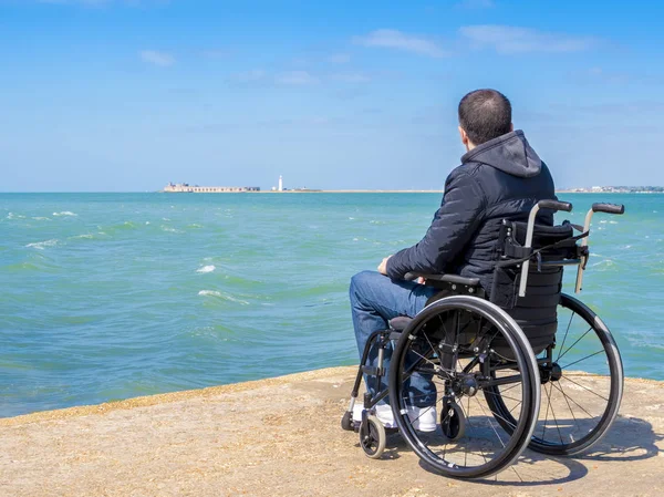Disabled young man sitting in a wheelchair and looks at the sea — Stock Photo, Image