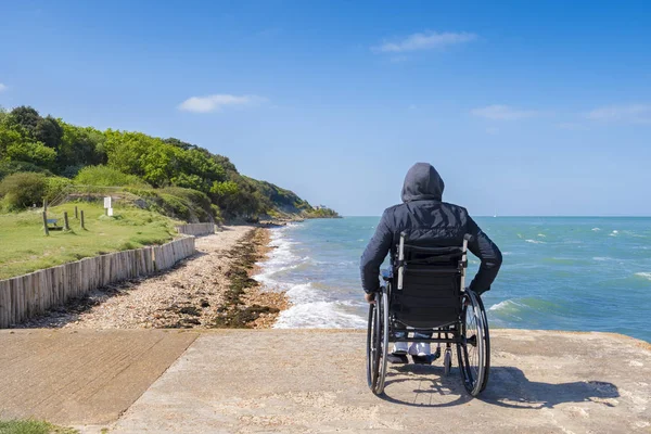 Jovem deficiente sentado em uma cadeira de rodas e olha para o mar — Fotografia de Stock