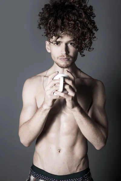 Attractive young man with long ginger curly hair having a cup of coffee or tea, studio portrait