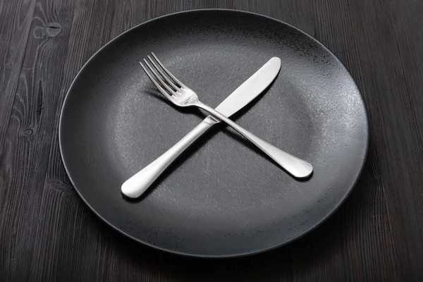 Black plate with crossing knife, spoon on dark — Stock Photo, Image