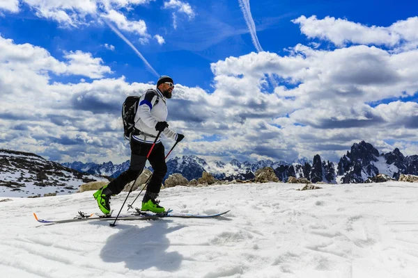 登山家野外滑雪春天走在白雪皑皑的山脊与滑雪板在背包里。在蓝色背景多云的天空和阳光在南蒂罗尔，白云岩，意大利蒙特优雅。冒险冬季极限运动. 免版税图库照片