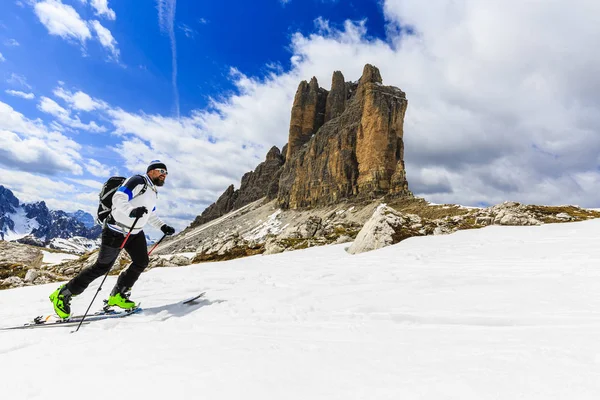 Bergsteiger-Backcountry-Skispringer, der mit Skiern im Rucksack auf einem schneebedeckten Grat bergauf geht. im Hintergrund blauer bewölkter Himmel und strahlende Sonne und monte cristallo in Südtirol, Dolomiten, Italien. Abenteuer Winter Extremsport. Stockbild