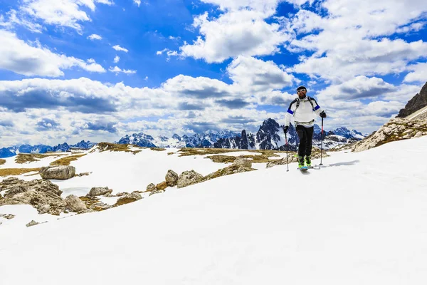 Bergsteiger-Backcountry-Skispringer, der mit Skiern im Rucksack auf einem schneebedeckten Grat bergauf geht. im Hintergrund blauer bewölkter Himmel und strahlende Sonne und monte cristallo in Südtirol, Dolomiten, Italien. Abenteuer Winter Extremsport. Stockbild