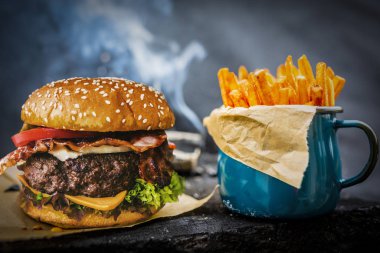 Lezzetli füme ızgara ve sırlı sığır eti burger marul, peynir ve domuz pastırması boşaltmak, duman mesquite ahşap ahşap arka plan ile ahşap masa üzerinde patates kızartması ile servis.