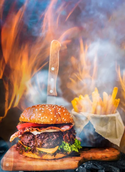 Tasty smoked grilled and glazed beef burger with lettuce, cheese and bacon served with french fries on wooden table with copyspace, smoke and fire wood in background.