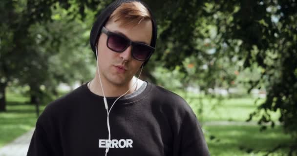 Mladý hipster poslouchá hudbu a usmívá se na kameru venku. — Stock video