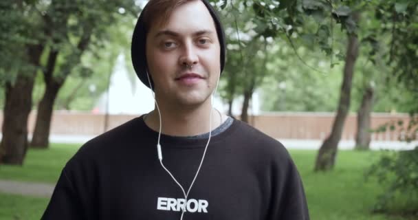 Joven hipster escucha música y sonríe en la cámara al aire libre . — Vídeos de Stock