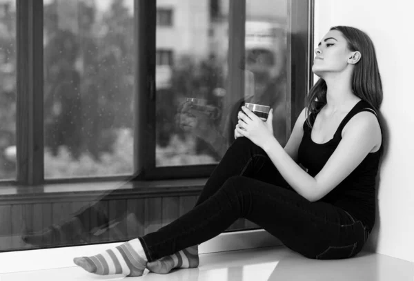 Young woman sitting with cup at windowsill at home in rainy day. — Stock Photo, Image