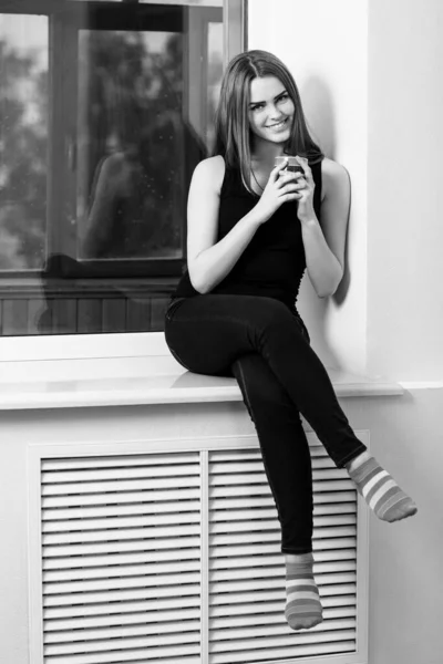 Ung kvinna sitter med kopp på fönsterbrädan hemma i regnig dag. — Stockfoto