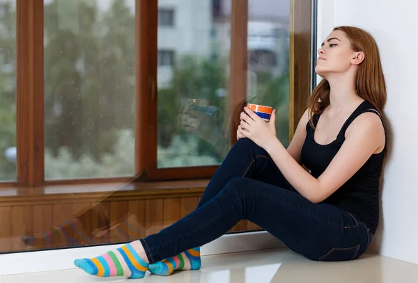 Mujer joven sentada con taza en el alféizar de la ventana en casa en días lluviosos . —  Fotos de Stock