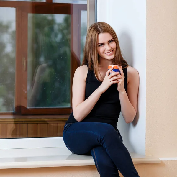 Mujer joven sentada con taza en el alféizar de la ventana en casa en días lluviosos . — Foto de Stock