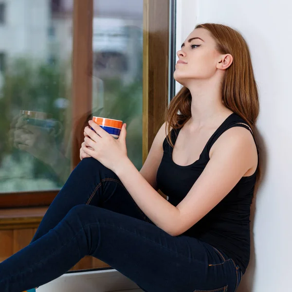 Young woman sitting with cup at windowsill at home in rainy day. — Stock Photo, Image