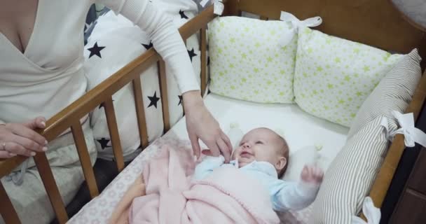 Madre sentada cerca de la cuna mira y toca a su bebé recién nacido . — Vídeos de Stock