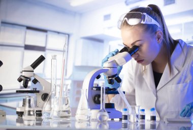 Bilim araştırma konsepti. Deney sırasında genç bir bilim adamı modern laboratuvarda mikroskop kullanıyor. Cam tüpler ve beherler..