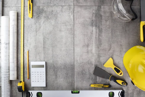Contractor concept. Tool kit of the contractor: yellow hardhat, libella, hand saw. Plans and notebook on the gray tiles background.