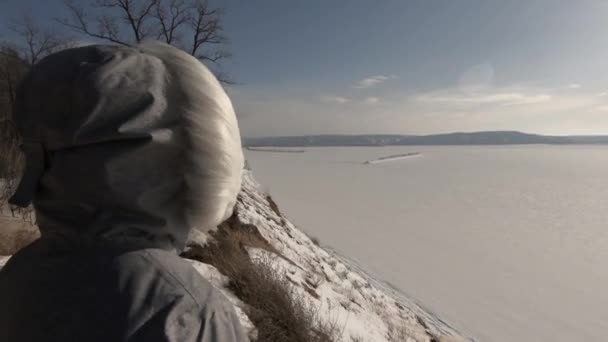 ジャケットを着た女性が崖の上に立っている 強い風と寒さ 広い川に氷と雪 フードと毛皮が見えます — ストック動画