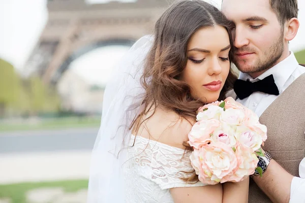 Mariée et marié assis sur le banc dans le parc, se tenant la main les uns des autres et bouquet. Groom tenant sa tête sur l'épaule de la mariée et la mariée souriant largement . — Photo
