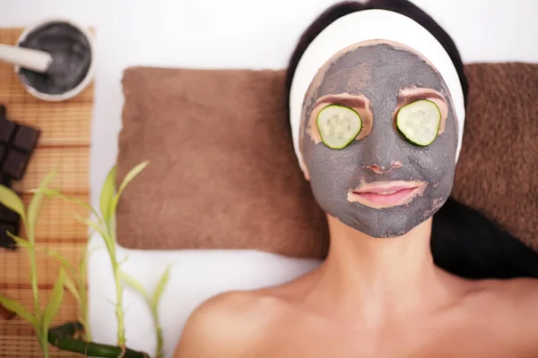 Beautiful young woman is getting facial clay mask at spa, lying with cucumbers on eyes — Stock Photo, Image