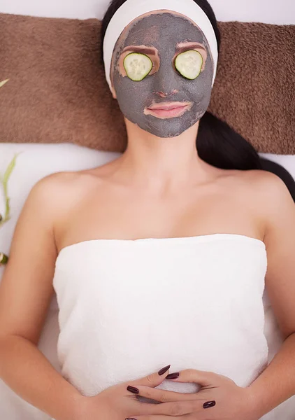 Hermosa joven mujer está recibiendo máscara de arcilla facial en el spa, acostado con pepinos en los ojos — Foto de Stock