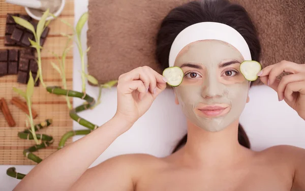 Hermosa joven mujer está recibiendo máscara de arcilla facial en el spa, acostado con pepinos en los ojos — Foto de Stock