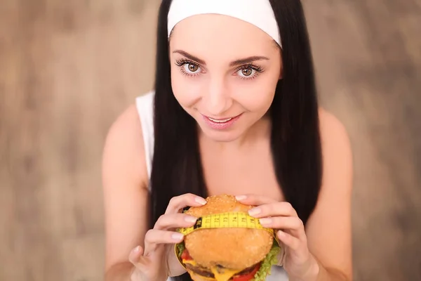 Une jeune femme tenant un hamburger dans un ruban à mesurer. Une fille se tient sur un fond en bois. La vue du haut. Le concept de saine alimentation . — Photo