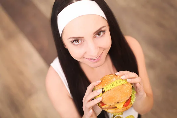 Eine junge Frau hält einen Burger in einem Maßband. steht ein Mädchen auf einem hölzernen Hintergrund. die Aussicht von oben. das Konzept der gesunden Ernährung. — Stockfoto