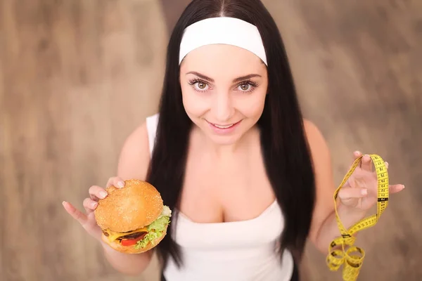 Eine junge Frau hält einen Burger und ein Maßband in der Hand. steht ein Mädchen auf einem hölzernen Hintergrund. die Aussicht von oben. das Konzept der gesunden Ernährung. — Stockfoto