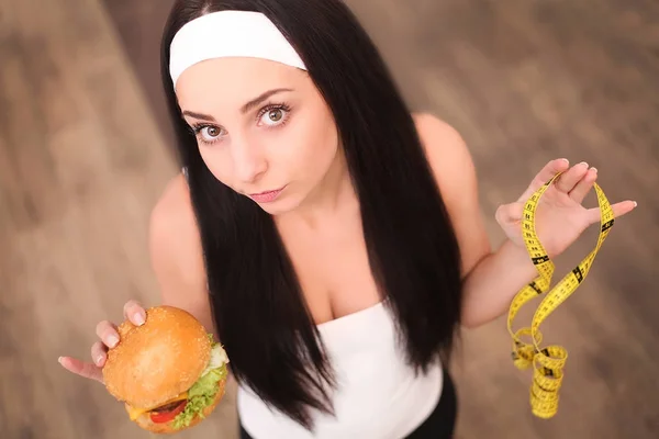 Eine junge Frau hält einen Burger und ein Maßband in der Hand. steht ein Mädchen auf einem hölzernen Hintergrund. die Aussicht von oben. das Konzept der gesunden Ernährung. — Stockfoto