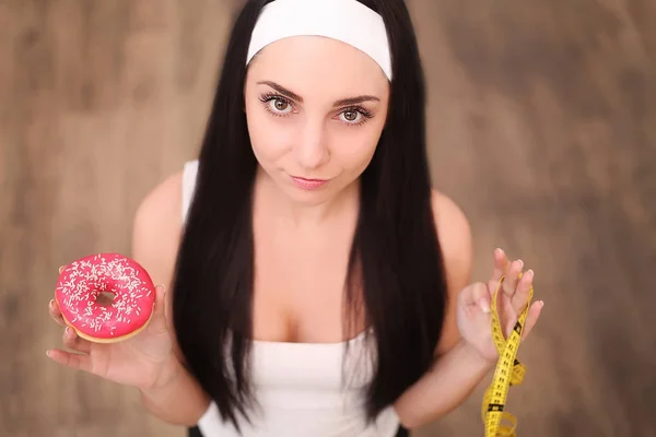 Una joven sosteniendo una rosquilla y una cinta métrica. Una chica está parada sobre un fondo de madera. La vista desde arriba. El concepto de alimentación saludable.Dieta . —  Fotos de Stock