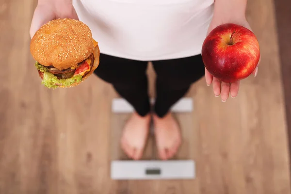 Dieta. Mujer Medición de peso corporal en báscula de pesaje Sosteniendo hamburguesa y manzana. Los dulces son comida chatarra malsana. Dieta, Comida Saludable, Estilo de Vida. La pérdida de peso. Obesidad. Vista superior — Foto de Stock