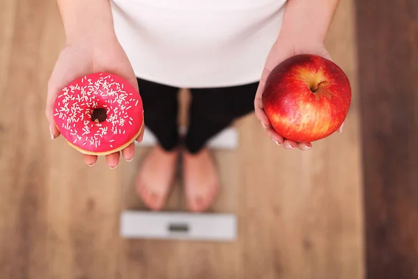 Δίαιτα. Γυναίκα μέτρησης σώματος βάρος για ζύγιση κλίμακας κρατώντας ντόνατ και μήλο. Τα γλυκά είναι ανθυγιεινά τρόφιμα παλιοπραγμάτων. Να κάνει δίαιτα, υγιεινή διατροφή, τον τρόπο ζωής. Απώλεια βάρους. Η παχυσαρκία. Το Top View — Φωτογραφία Αρχείου