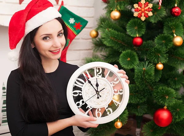 Feliz joven mujer está sosteniendo un reloj y sonriendo mientras celebra la Navidad en casa —  Fotos de Stock