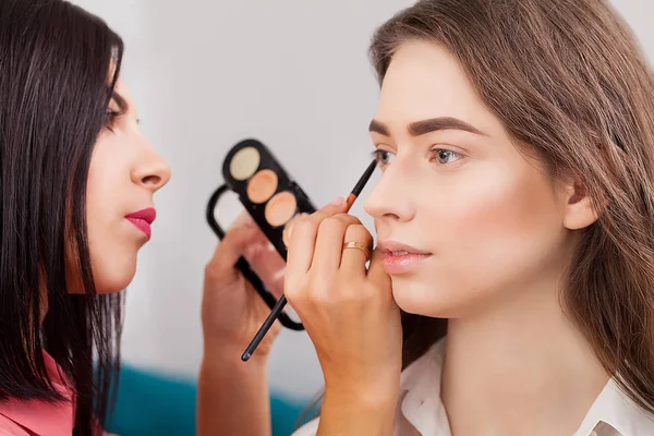 Maskér dělá profesionální make-up mladé ženy u zrcadla v beauty studio — Stock fotografie