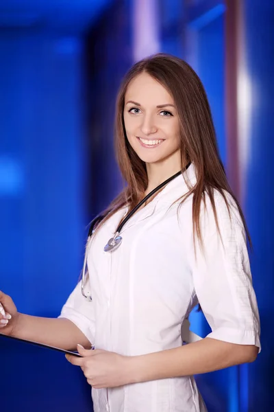 Medicina. Uma médica com uma pasta no hospital. Estudante graduado. Menina jovem e bonita. Roupão branco. No fundo azul do hospital. No pescoço é um estetoscópio . — Fotografia de Stock