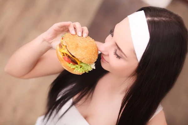 Unhealthy eating. Junk food concept. Portrait of fashionable young woman holding burger and posing over wood background. Close up. Copy-space. Perfect hair, skin, make-up and manicure. Studio shot