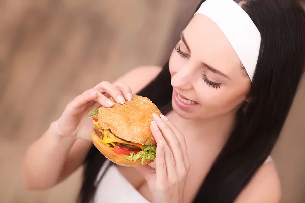 Comida pouco saudável. Conceito de comida de plástico. Retrato de mulher jovem na moda segurando hambúrguer e posando sobre fundo de madeira. Fecha. Espaço de cópia. Cabelo perfeito, pele, maquiagem e manicure. Estúdio — Fotografia de Stock