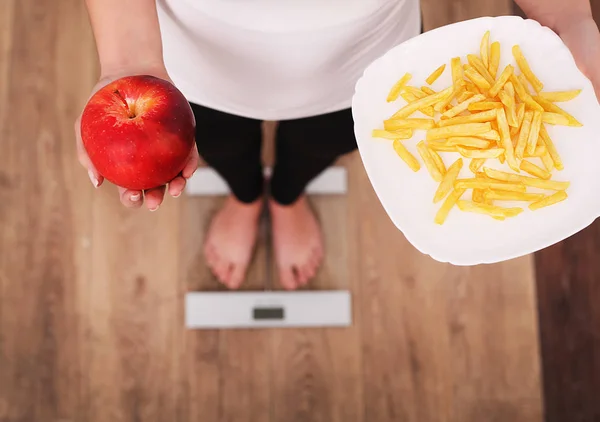 Молодая красивая женщина стоит на весах и делает выбор между яблоком и картофельными чипсами. Понятие здорового питания. Здоровый образ жизни. Диета . — стоковое фото