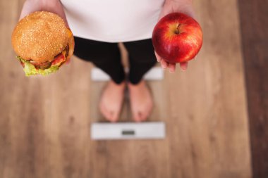 Diyet. Kadın ölçme vücut ağırlığı üzerinde ağırlığında ölçek Holding Burger ve elma. Tatlılar sağlıksız abur cubur var. Diyet, sağlıklı beslenme, yaşam tarzı. Kilo kaybı. Obezite. Üstten Görünüm