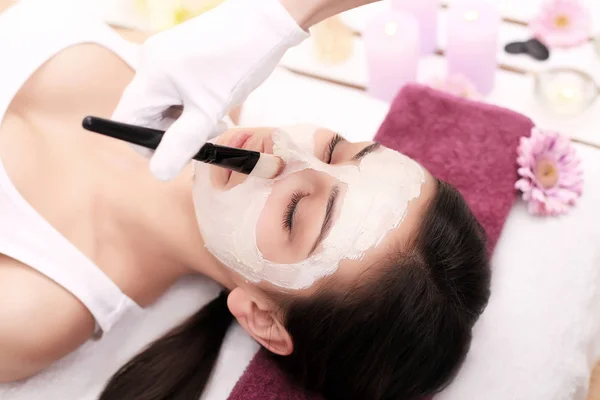 Hermosa joven mujer está recibiendo máscara de arcilla facial en el spa, mintiendo —  Fotos de Stock