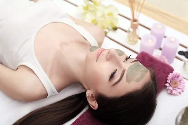 Beautiful young woman is getting facial clay mask at spa, lying — Stock Photo, Image